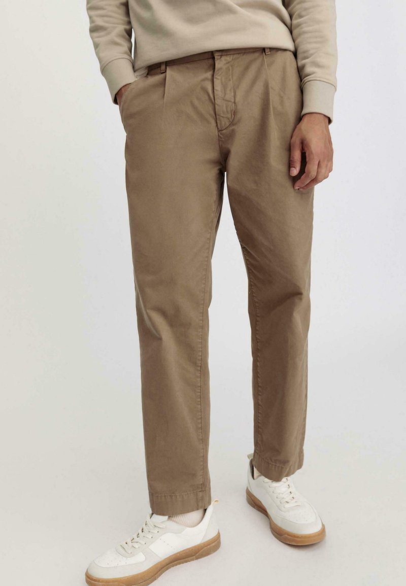 Pantaloni di cotone color sabbia con vestibilità rilassata, frontale plissettato e tasche laterali, abbinati a sneaker bianche con suola in gomma.