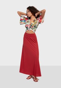 Květinový crop top s zvonovými rukávy v živých barvách, spárovaný s dlouhou, jednobarevnou červenou sukní vyrobenou z hladké látky. Celý vzhled doplňují jednoduché červené sandály.