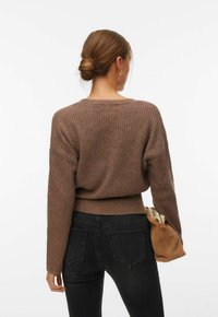 Femme aux cheveux bruns attachés en chignon, portant un pull marron côtelé, un jean noir, et tenant une pochette en daim beige sur un fond blanc.