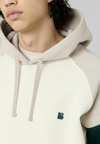 Sweat-shirt à capuche crème avec des accents beiges, col rond et logo imprimé vert sur la poitrine, doté de cordons de serrage et d'une texture douce.