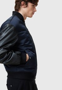 Navy bomber jacket with black leather sleeves, ribbed cuffs and hem, zip closure, and side pockets. Smooth and shiny texture.