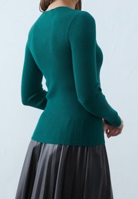 Femme aux longs cheveux portant un pull côtelé ajusté couleur sarcelle et une jupe plissée foncée, vue de dos sur un fond uni.