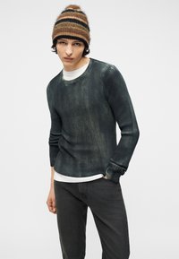 Νεαρός άνδρας που φοράει καφέ ριγέ σκουφί, σκούρο πράσινο ριμπ πλεκτό πουλόβερ πάνω από λευκό πουκάμισο και μαύρα τζιν, με το ένα χέρι στην τσέπη.