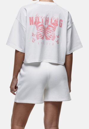 Person trägt ein weißes, kurz geschnittenes T-Shirt mit einem pinkfarbenen Schmetterling und dem Schriftzug „Good For Nothing Atelier“, kombiniert mit weißen Shorts, Ansicht von hinten.