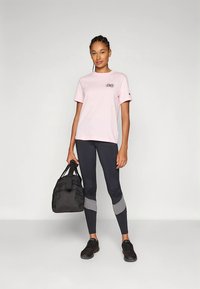 T-shirt rosa curta com um logótipo, combinada com leggings escuras com detalhes cinzentos. A modelo segura uma mala de ginásio preta e usa sapatilhas escuras.
