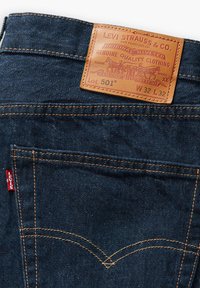 Jeans de mezclilla azul oscuro con un parche de cuero que muestra "Levi Strauss & Co.", costuras naranjas y una etiqueta roja cerca del bolsillo trasero.