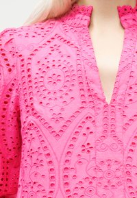 Φωτεινή ροζ μπλούζα με περίτεχνη κεντητή δαντέλα και γιακά με κυματιστό τελείωμα, φορεμένη από άτομο με μακριά ξανθά μαλλιά.