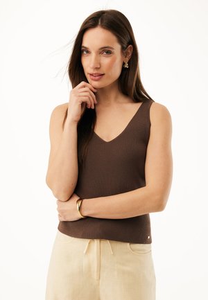 Femme aux longs cheveux bruns portant un haut marron sans manches, un pantalon beige, un bracelet en or et des boucles d'oreilles, posant avec la main près du visage sur fond blanc.