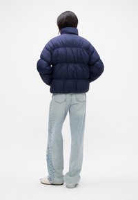 Granatowa puchowa kurtka z poziomymi przeszyciami, wysokim kołnierzem i szerokimi rękawami. Noszona z jasnoniebieskimi szerokimi dżinsami i jasnymi sneakersami.