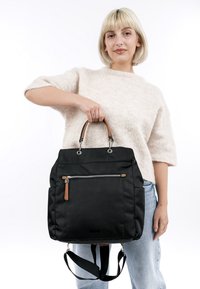 Woman with short blonde hair wearing a beige sweater and light jeans holds a large black handbag with brown handles and a front zipper pocket.