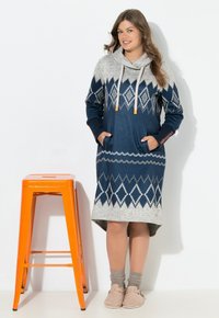 Glunderende vrouw in een blauw-grijs gestreepte trui-jurk en beige slippers staat naast een hoge oranje metalen kruk tegen een witte muur.