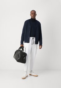 Ναυτικό πλεκτό ζακέτα πάνω από μαύρο πουλόβερ, λευκά cargo παντελόνια με μαύρη κορδόνι, κρατώντας μια μαύρη δερμάτινη τσάντα duffle, λευκά αθλητικά παπούτσια.