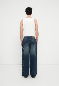 Široke plave traperice kroja širokih nogavica s izblijedjelim završetkom, izvezeni dizajn na lijevoj nozi i klasični džepovi straga. Uparene s bijelom majicom bez rukava.