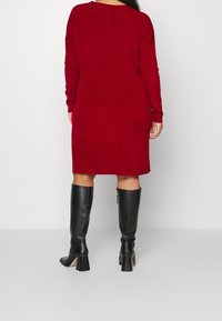 Robe rouge en maille côtelée avec manches longues, encolure ronde et ourlet droit. Associée à des bottes en cuir noir à talons montant jusqu'au genou.