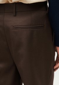 Gros plan sur un pantalon marron sur mesure avec une poche passepoilée horizontale à l'arrière, porté avec un haut beige et une manche bleu marine visible.
