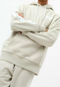 Personne portant un sweat à capuche beige et un pantalon de jogging assorti, les bras croisés, cordons blancs visibles et logo discret sur la manche et la poitrine.