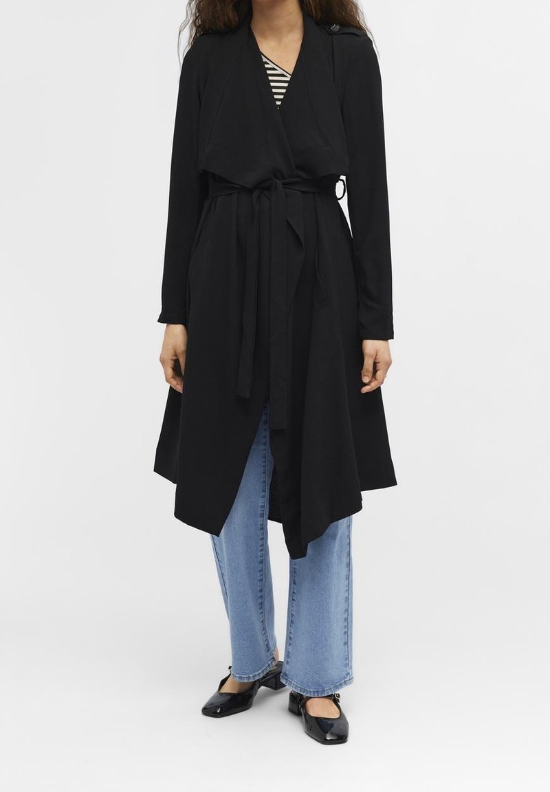 Veste longue noire avec taille ceinturée, devant ouvert et manches longues, associée à un haut rayé et un jean bleu coupe large. Texture lisse.