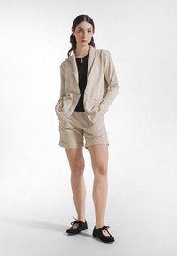 Blazer beige con un solo bottone e due tasche, abbinato a pantaloncini beige con risvolti arrotolati e scarpe nere stringate.