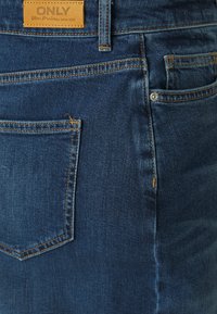 Jeans de denim azul-escuro com uma textura suave, costura laranja, um bolso traseiro e um patch de logotipo em couro na cinta.