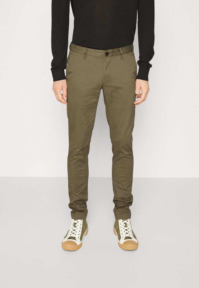 Zelené slim fit kalhoty vyrobené z bavlny s knoflíkovým zapínáním, předními kapsami a kombinované s bílými teniskami s hnědými podrážkami.