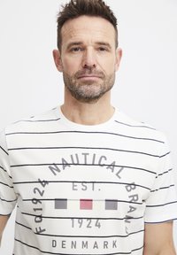 Baltos medvilnės marškinėliai su tamsiai mėlynomis horizontaliomis juostelėmis, apvaliu kaklu ir grafikiniu piešiniu su datomis ir spalvotais blokais priekyje.