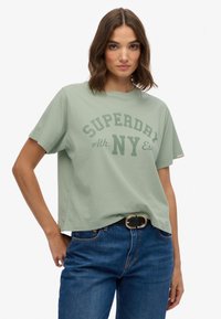 Camiseta de algodón verde claro con un gráfico de "SUPERDRY" en verde oscuro, mangas cortas, corte relajado, combinada con jeans azules y un cinturón negro.