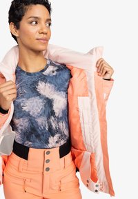 Waterdichte roze jas met een zachte voering, gecombineerd met een blauw shirt met abstract patroon en perzikkleurige broek met ritsen en knoopaccenten.