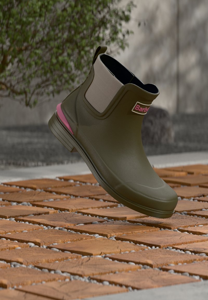 Green rubber ankle boot with light gray elastic sides and a pink accent on the sole, displayed above a wooden surface with pebbles.