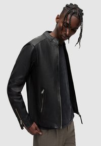 Schwarze Lederjacke mit Stehkragen, Reißverschluss vorn und zwei seitlichen Taschen. Strukturiertes Finish mit enganliegenden Ärmeln und silbernen Hardware-Akzenten.