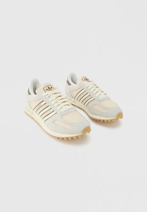 Ζευγάρι Μπεζ και Λευκά αθλητικά παπούτσια Adidas LA Trainer με καουτσούκ σόλες και μαύρες ρίγες, ελαφρώς γερμένα προς τα δεξιά πάνω σε λευκό φόντο.