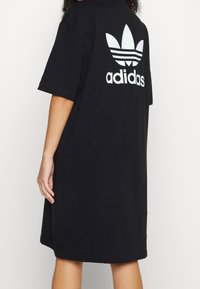 Černé oversized polo šaty vyrobené z bavlny, s bílým logem Adidas a designem trojlístku na zadní straně. Krátké rukávy a volný střih.