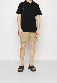 Polo noir, short beige et sandales foncées. Matière en coton, manches courtes, design uni, et chaussures à semelle plate avec brides croisées.