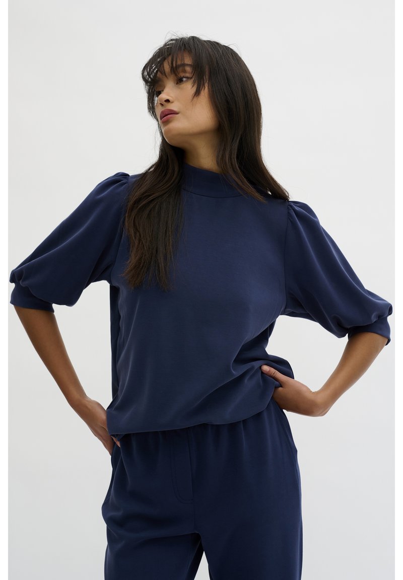 Navy blue, loose-fitting top with a high collar and puffed sleeves, made of smooth fabric. Worn with matching pants, minimal texture.