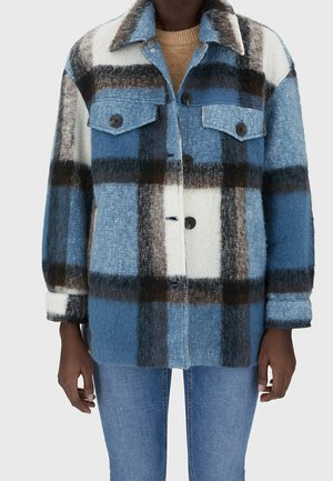 Personne porte une veste en laine à carreaux bleue, blanche et marron boutonnée par-dessus un pull beige et un jean bleu, debout avec les bras détendus.