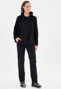 Sort softshell-jakke med hætte, parret med sorte bukser, begge designet til udendørs aktiviteter. Har lynlåse og lommer.