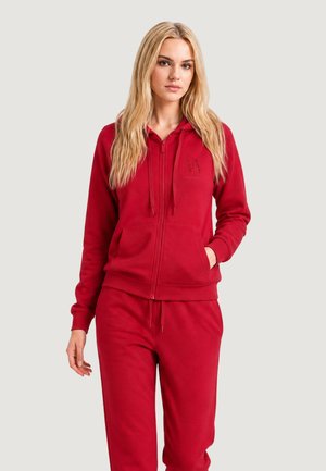 Femme blonde portant un sweat à capuche rouge zippé et un pantalon de jogging rouge assorti, debout avec une main dans la poche devant un fond uni.