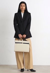 Zwarte op maat gemaakte blazer, witte blouse, beige wijde broek, houdend een gestructureerde crème handtas met zwarte accenten en een gouden sluiting.