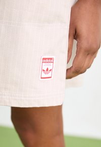 Lähenemne käsi beeži triibuliste lühikeste pükste kõrval, mille alläärtes on punane ja valge Adidas'i logo plekk.