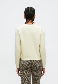 Pull crème à finition texturée, col rond et manches longues, associé à un pantalon à motifs rayés marron et beige.