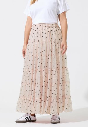 Person iført en beige, lagdelt, prikket maxi-nederdel kombineret med en hvid t-shirt og hvide sneakers med sorte striber.