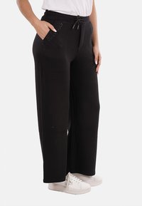 Persona che indossa pantaloni larghi neri con vita regolabile e scarpe da ginnastica bianche, in piedi con una mano in tasca e l'altra al fianco.