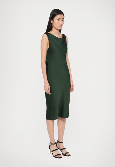 Zielona sukienka bez rękawów z satyny o dopasowanym kroju, długości do kolan i gładkiej fakturze. Noszona z czarnymi sandałami na wysokim obcasie ze paskami.