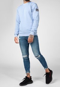 Hombre con sudadera azul claro con un parche que dice "Amigos Equivocados", jeans ajustados rasgados, zapatillas negras con texto rojo y tatuajes visibles en las manos.