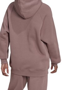 Przesadna bluza z miękkiego materiału w kolorze intensywnego różu, z kapturem, raglanowymi rękawami i ściągaczami. Tył ma gładki, minimalistyczny design.