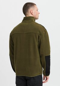 Olivegrønn fleecepull med høy krage, svarte ermer med detaljer og sømmer; teksturert stoff med en avslappet passform.