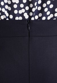 Marinblå kjol med en strukturerad yta, med en svart dragkedja bak. Ovandelen har ett prickigt mönster i vitt på marinblått.