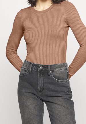 Kvinde iført en tætsiddende, ribstrikket, brun bluse med lange ærmer, stoppet ned i højtsiddende, falmede sorte jeans med hænderne bag ryggen.