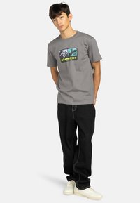 Grauema Baumwoll-T-Shirt mit einem grafischen Design aus Bergen und Eidechsenmustern, kombiniert mit schwarzen Jeans und weißen Turnschuhen.
