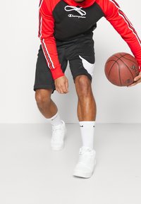 Pantaloni neri con accenti bianchi, felpa rossa a maniche lunghe, sneaker bianchi e pallone da basket. Tessuto testurizzato, logo visibile sulla felpa.