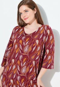 Tunic bordeaux con un motivo paisley in bianco e oro. Presenta un collo rotondo e maniche a tre quarti. Tessuto morbido.